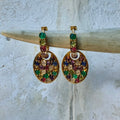 Gold earrings with multicolored gemstones on a wooden stick against a textured white background