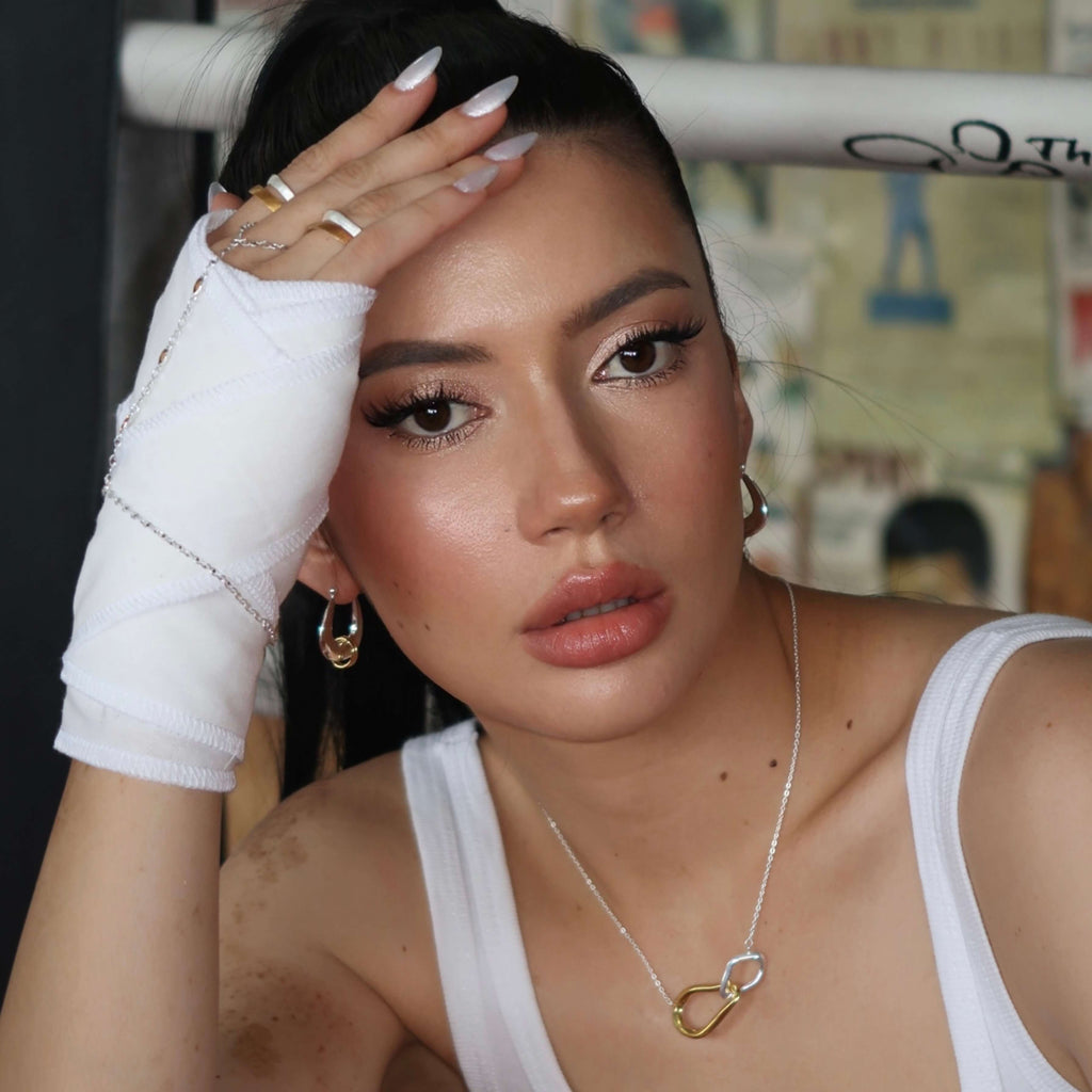 Woman with white boxing gloves and bandages, wearing a mixed metal hoop earrings and a heart-shaped necklace, in a casual setting.