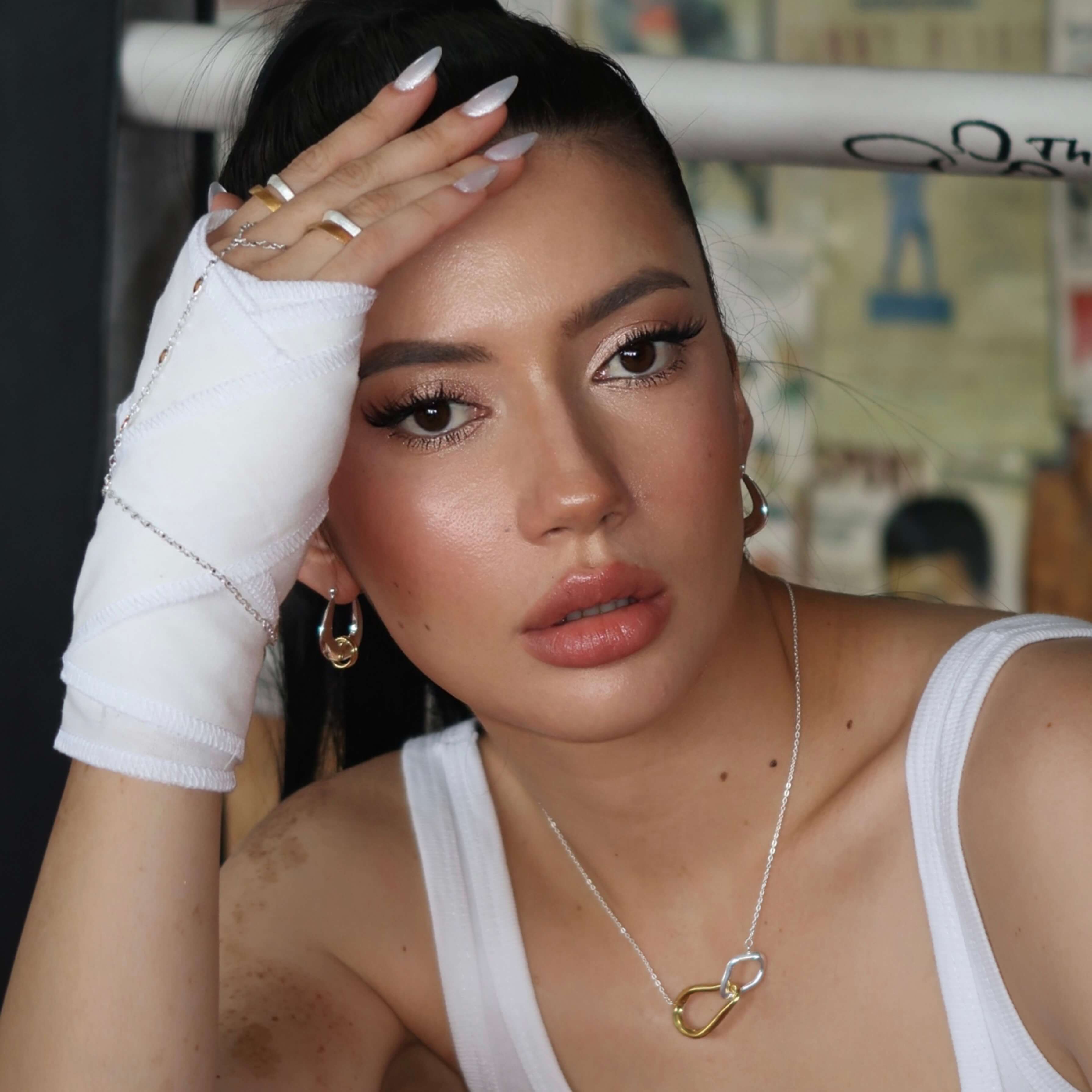 Woman with white boxing gloves and bandages, wearing a mixed metal hoop earrings and a heart-shaped necklace, in a casual setting.