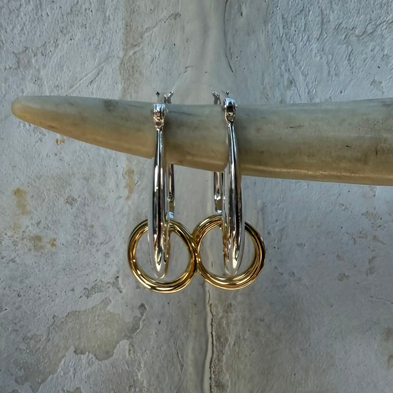Gold hoop earrings hanging on a wooden stick against a textured wall.